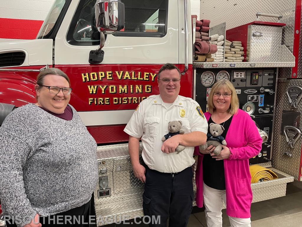Chief Justin Lee, President of the Southern League poses with Margaret Teehan (left) and Lori Roeleveld (right) in front of Hope Valley Tanker 915 along with one of the teddy bears donated by TriCounty Community Action to the League's Departments. 